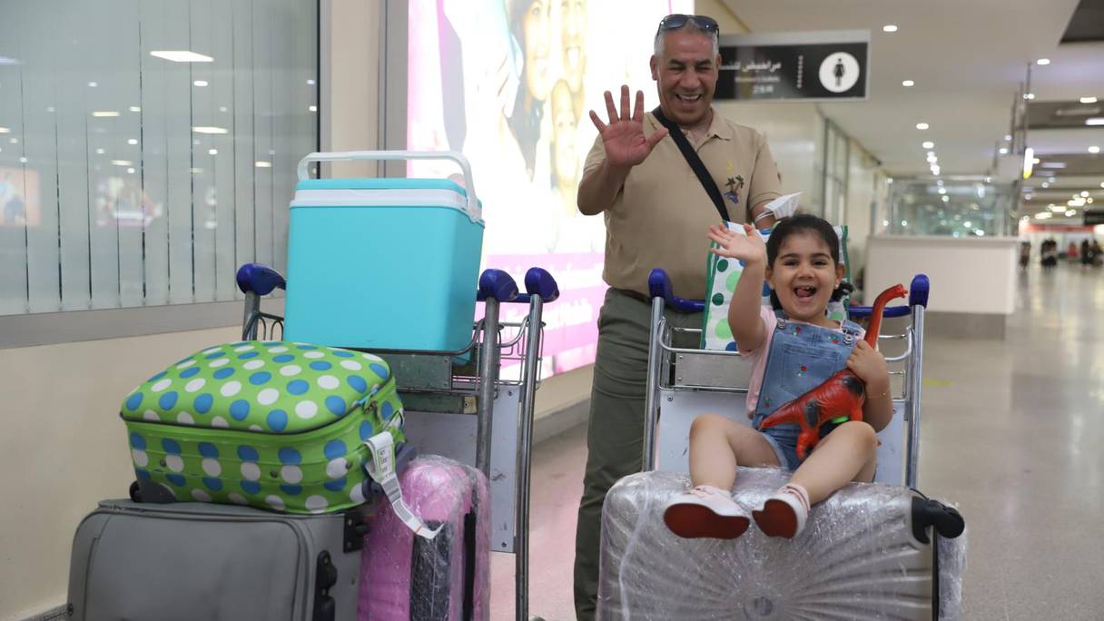 Une petite fille et un homme, Marocains résidant à l’étranger, arrivés à l’aéroport Casablanca-Mohammed V, font signe à la caméra en souriant.
