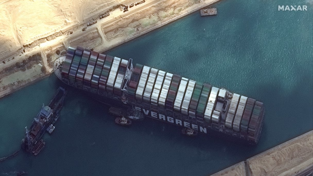 Image satellite où des remorqueurs et des dragues tentent de libérer le cargo Evergreen le 26 mars 2021, dans le canal de Suez. Le porte-conteneurs, équivalent de plus de quatre terrains de football, est calé en diagonale sur tout le canal depuis le 23 mars, fermant la voie navigable dans les deux sens. 
