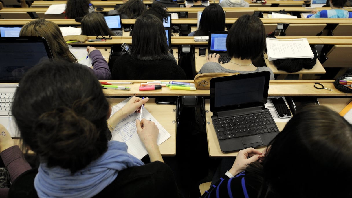 Des étudiants en cours magistral, à leur pupitre, dans un amphithéâtre d'université. 
