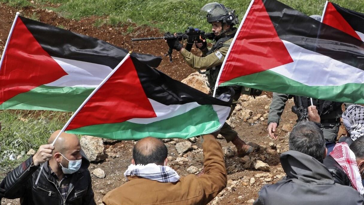 Des manifestants palestiniens agitent leur drapeau alors qu'un membre des troupes israéliennes pointe son arme, lors d'une manifestation contre l'établissement d'avant-postes israéliens sur leurs terres, à Beit Dajan, à l'est de Naplouse en Cisjordanie occupée, le 2 avril 2021.
