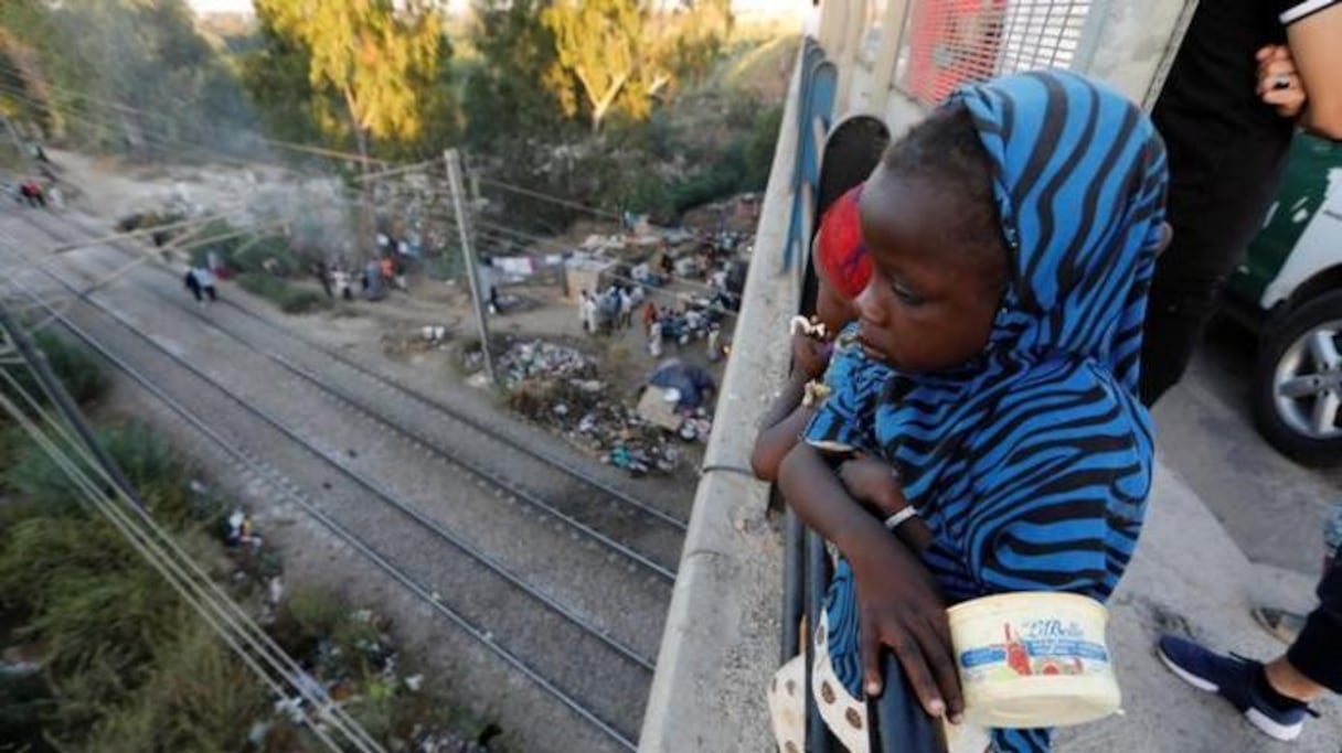 Une jeune migrante sur un pont routier surplombant un camp de fortune.
